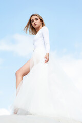 Fototapeta na wymiar Beautiful young blonde woman bride in white dress outdoor on the sand and blue sky background