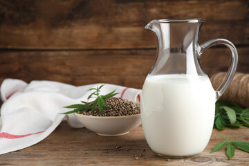 Glass jug of fresh hemp milk, seeds and leaves on wooden table. Space for text