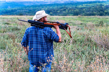 A man with a hat and a rifle on his shoulder stands with his back to the camera and looks to the...