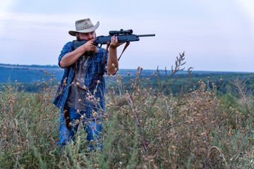 Fototapeta premium A hunter with a beard and hat shoots a rifle in the field, there is little noise, soft focus