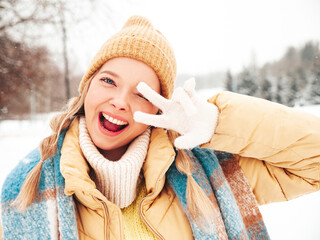 Young beautiful smiling hipster female in trendy warm clothes and scarf.Carefree woman posing in the street in park. Positive pure model having fun in snow. Enjoying winter moments. Shows peace sign