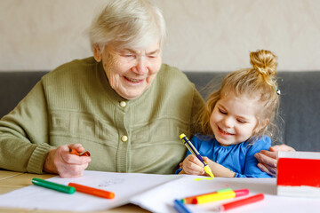 Beautiful toddler girl and grand grandmother drawing together pictures with felt pens at home. Cute...