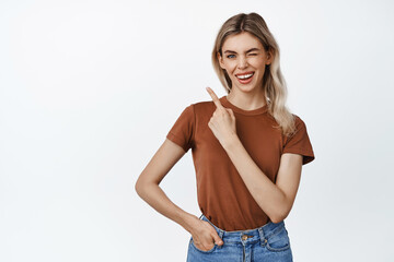 Smiling blond girl winking at you, pointing finger at upper left corner sale advertisement, showing...