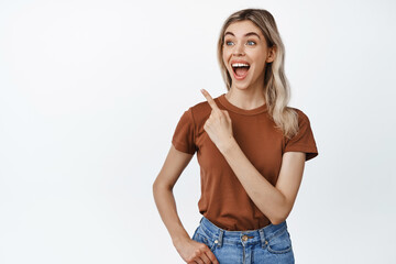 Fototapeta premium Young happy girl watching smth funny, pointing finger at upper left corner and looking amazed, laughing out loud, standing over white background