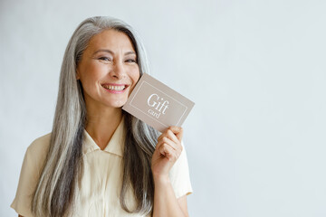 Pretty smiling mature Asian lady holds gift card posing on light grey background in studio, space...