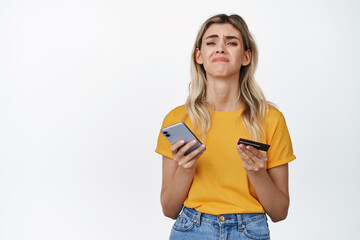 Upset girl holding smartphone and credit card, concept of woman lacking money, cant buy something, standing over white background