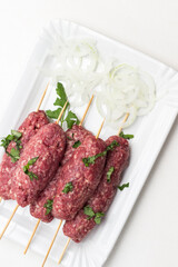 Raw minced meat on wooden skewers and chopped onion rings on white plate.