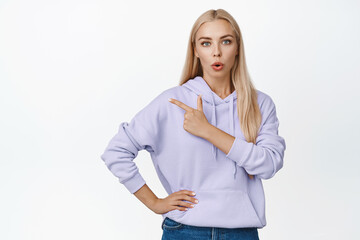 Curious blond woman asks question about product, pointing finger left and looking surprised, standing over white background