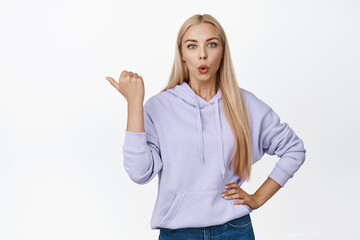 Surprised blond woman pointing finger left, gasping and looking impressed, recommending to check out something interesting, standing over white background