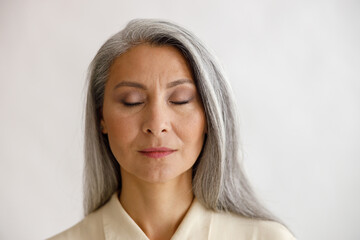 Pretty tranquil grey haired lady with closed eyes and elegant makeup poses on light background in...