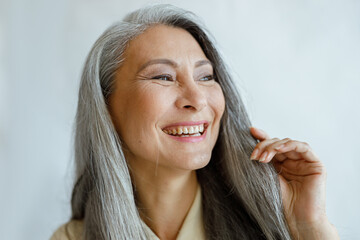 Joyful middle aged Asian woman with natural grey hair smiles on light background in studio closeup....