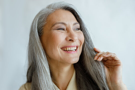 Joyful Middle Aged Asian Woman With Natural Grey Hair Smiles On Light Background In Studio Closeup. Mature Beauty Lifestyle