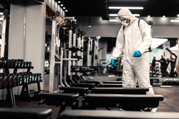 Disinfecting the air, space, and gym equipment. A man in white protective clothing and mask cleans...