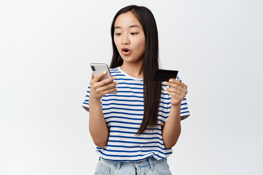 Image of asian girl holding credit card, looks excited at mobile phone screen, paying for online shopping, using store app, standing over white background