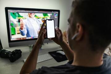 A man holding a smart phone with a white screen in front of a desktop screen and a photo camera in the office. Free space for advertising and marketing messages. Home office atmosphere