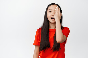 Image of asian girl covers one side of face, looks surprised and gasping startled, stands against white background