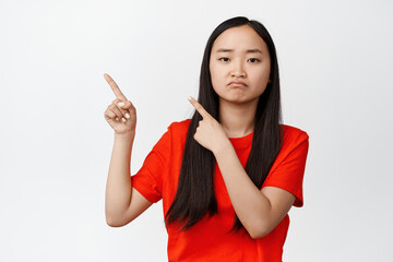 Sad and moody asian woman pointing fingers at upper left corner, pouting and frowning upset, standing in red t-shirt against white background