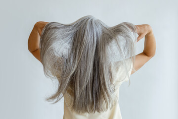 Middle aged woman in yellow blouse shows long loose grey hair standing on light background in studio backside view. Mature beauty lifestyle