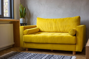 Bright yellow sofa stands in the room near the window and grey wall. Modern interior design, loft style