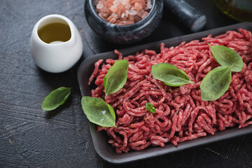Close-up of raw beef mincemeat with fresh green basil, pink salt and olive oil, studio shot on a dark-brown stone surface