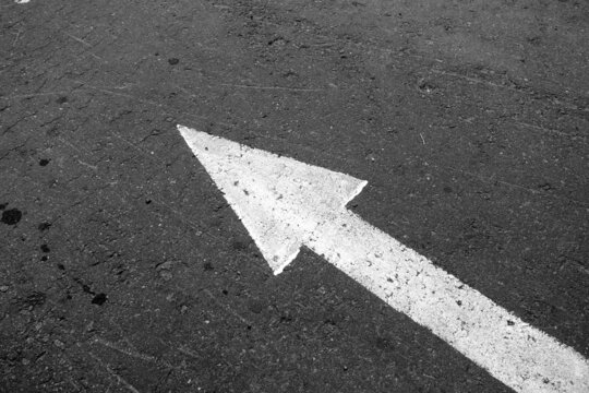White arrow on asphalt pointing left in black and white.