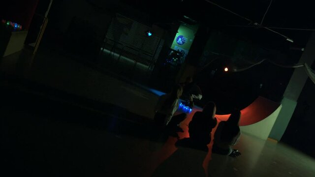 Young guy doing stunt on ramp roller-skating. Millennial showing off with girls watching in dark studio. Teenager making trick on glowing wheels. Concept of lifestyle, activity, street culture.