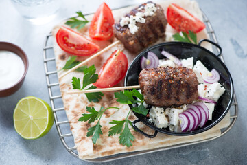 Grilled kofta kebabs with flatbread, feta cheese and tomatoes, studio shot on a light-blue stone background, selective focus