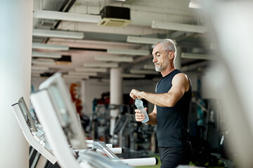 Mature athletic man drinks water after running on treadmill in gym.