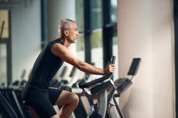 Fototapeta na wymiar Mature sportsman uses exercise bike while working out in gym.