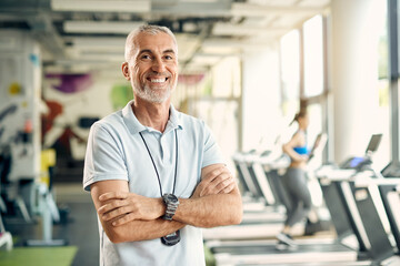 Happy mature personal trainer stands with arms crossed in gym and looks at camera.