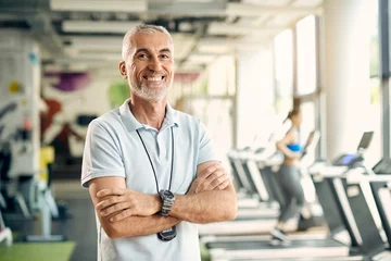  Happy mature personal trainer stands with arms crossed in gym and looks at camera. © Drazen