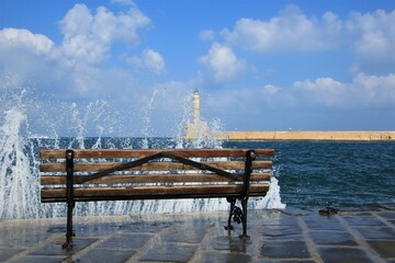 Sharp waves at the harbor edge with blue sky, lighthouse in the background and a bench in the...