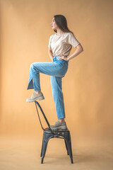 Teen girl in denim jeans and casual 90s clothes standing on old chair in studio. Beige background. Natural posing and stylish design photo