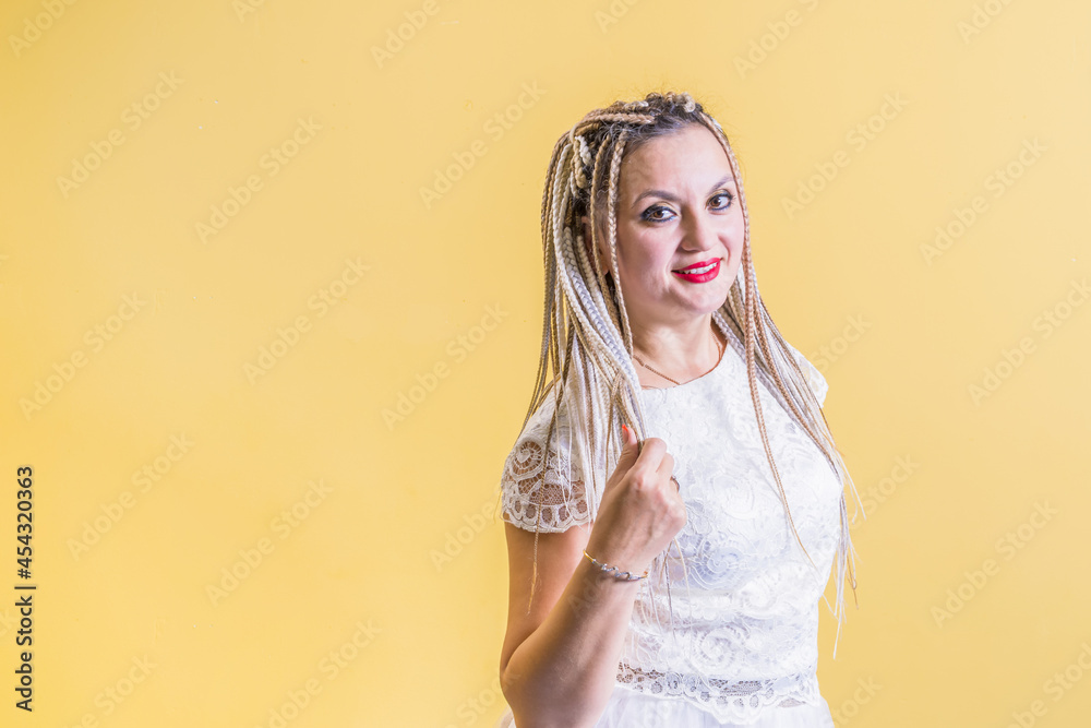 Poster Happy woman with African braids in white dress stands on yellow background. Looks into camera.