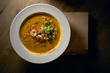 Creamy orange pumpkin soup with shrimp and spring onion photographed from above. The pumpkin puree is served in a large white dish on a wooden table with soft light. Homemade orange vegetable cream.