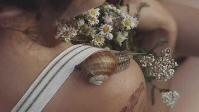 Close-up of snail slowly crawling along shoulder of young girl. Woman holding bouquet of wild flowers or daisies in her hands. Snail leaves muscus on skin. Concept of nature, cosmetology, spa, beauty.