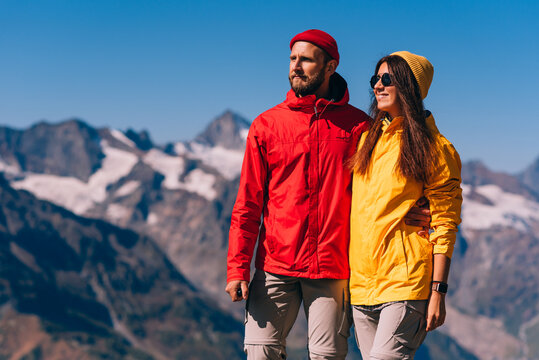 A traveling couple in hiking gear against the background of mountains. Two tourists on the top of the mountain. A man and a woman in the mountains. Two travelers in colorful jackets, close-up.