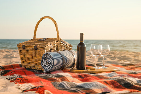 Blanket with picnic basket, corkscrew, bottle of wine and glasses on sandy beach near sea