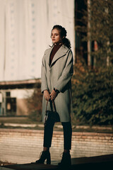 Young fashionable beautiful girl in a long gray coat, black jeans and with a dark red bag. Portrait shooting of a stylish girl. Facial shooting, street style and fashion. Professional model.