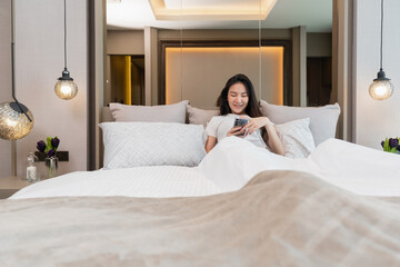 Bedroom concept a girl with drowsy feeling laying her back on several big and comfortable pillows on the bed looking on the mobile screen for social media