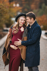 Young stylish couple in love at an autumn city park. Happy stylish man and beautiful pregnant woman in modish dress and hat posing outdoors on an autumn day