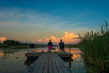 Couple man and woman seated on a wooden jetty, looking a colorful sunset on the lake and fishing