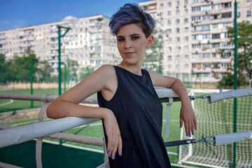 Stylish millennial girl with purple colored hair wearing little black dress at the school stadium...