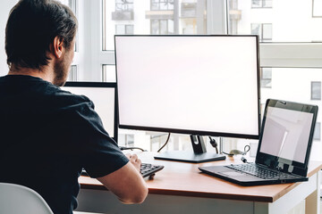 Man sitting at workplace with two laptops and monitor near the window. Remote work from home