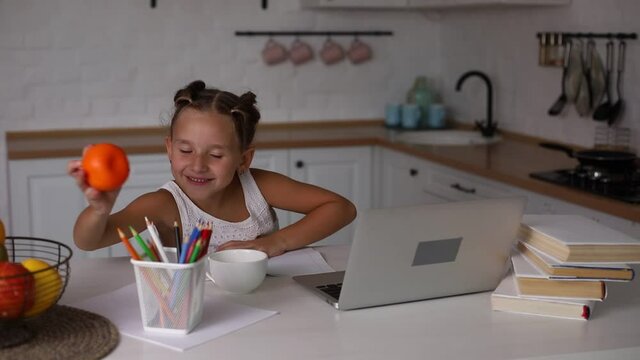 Little pretty girl with two ponytails sitting at kitchen at table with exercise book and laptop doing her school lessons because of lockdown quarantine. Home family education concept.