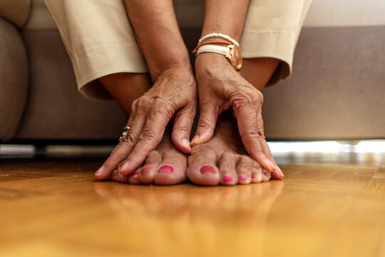 Close up of senior woman hands touching legs with varicose veins, sitting on sofa at home. Woman suffering from ankle pain. An older woman massages her feet to relieve the pain caused by rheumatism.