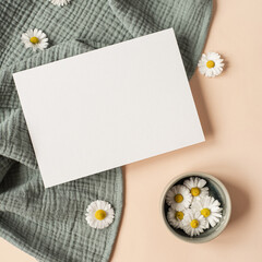 Obraz premium Flatlay aesthetic Parisian styled invitation template. Blank paper sheet card with mockup copy space, chamomile flower buds and aquamarine muslin cloth on neutral beige background. Flat lay, top view