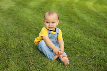 a beautiful little boy in a yellow T-shirt and a denim suit is sitting on the lawn in the park
