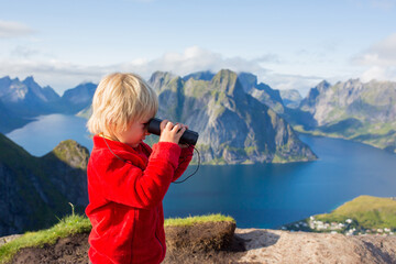 Cute child, standing on top of the mountains and looking down on Reine after climbing Reinebringen treeking path with lots of stairs, using binoculars