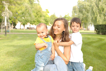 a young beautiful mother hugs
her young sons sitting on the lawn in the park